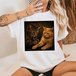 May include: White t-shirt featuring a square image of two cats. One cat is tattooing a fishbone on the other cat's arm. The cat doing the tattoo is brown and the cat getting the tattoo is orange. The image has a dark background.