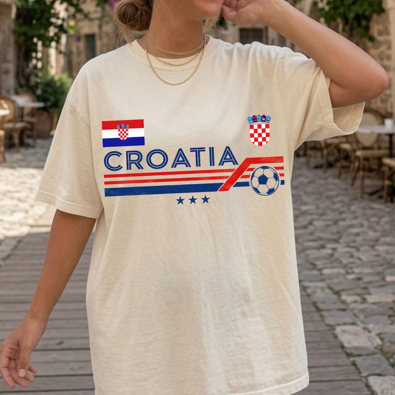 May include: A cream-colored short-sleeve t-shirt featuring the text CROATIA in blue, a red, white, and blue flag, a red and white checkered crest, a soccer ball graphic, and three blue stars arranged in a horizontal line across the chest.