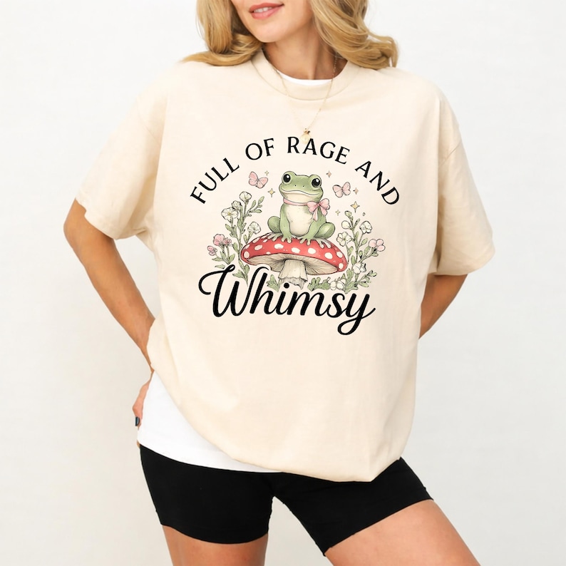 Puede incluir: Camiseta color crema con el texto "FULL OF RAGE AND WHIMSY". El gr&aacute;fico presenta una rana verde con pajarita, sentada sobre una seta roja y blanca, rodeada de flores y mariposas. La camiseta tiene mangas cortas.