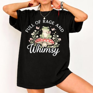 Puede incluir: Camiseta negra con un gr&aacute;fico caprichoso. El dise&ntilde;o presenta una rana con un lazo rosa, sentada en una seta roja y blanca, rodeada de flores y mariposas. El texto dice "FULL OF RAGE AND Whimsy."