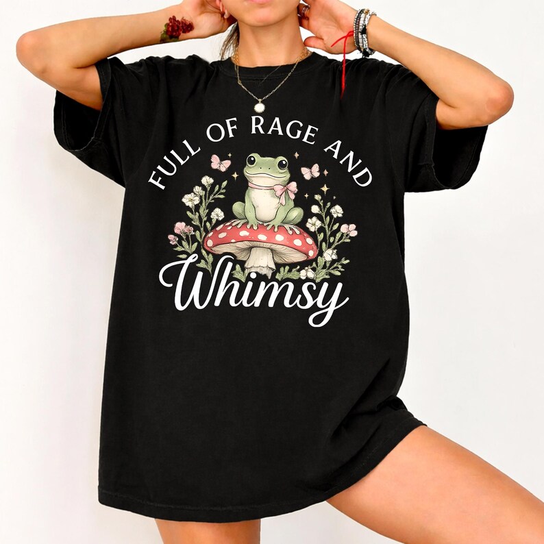 Puede incluir: Camiseta negra con un gr&aacute;fico caprichoso. El dise&ntilde;o presenta una rana con un lazo rosa, sentada en una seta roja y blanca, rodeada de flores y mariposas. El texto dice "FULL OF RAGE AND Whimsy."