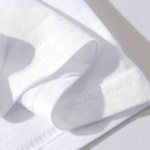 Puede incluir: Primer plano de una manga de camiseta blanca, que muestra la tela doblada y el dobladillo cosido. La imagen destaca la textura y la construcci&oacute;n de la prenda, con costuras visibles y una est&eacute;tica limpia y minimalista. El color blanco brillante sugiere simplicidad y versatilidad.