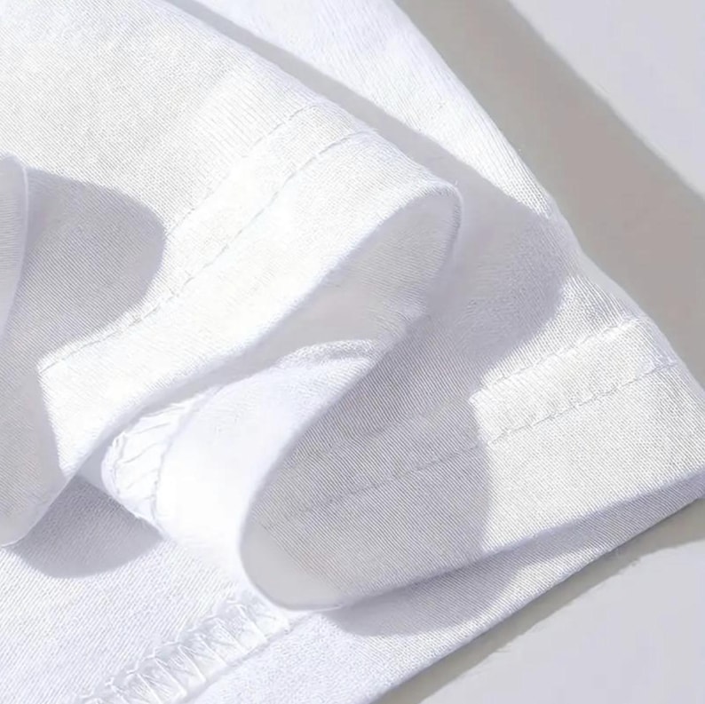 Puede incluir: Primer plano de una manga de camiseta blanca, que muestra la tela doblada y el dobladillo cosido. La imagen destaca la textura y la construcci&oacute;n de la prenda, con costuras visibles y una est&eacute;tica limpia y minimalista. El color blanco brillante sugiere simplicidad y versatilidad.