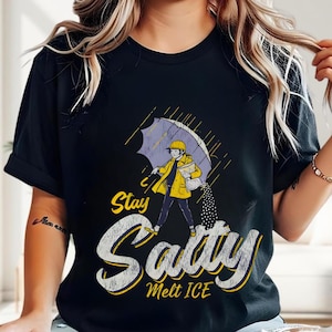 May include: Black t-shirt with a graphic of a person in a yellow raincoat and hat under an umbrella. The text "Stay Salty Melt ICE" is in a distressed font. The design features rain and a person walking.