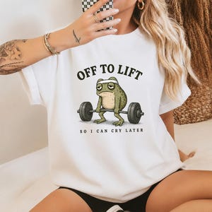 May include: White t-shirt with the text "OFF TO LIFT" above a cartoon frog lifting weights. The frog has a white headband and a sad expression. The text "SO I CAN CRY LATER" is below the frog.