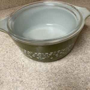 May include: An olive green Pyrex casserole dish with a clear glass lid. The dish has white floral patterns around the base and two handles. The interior is a light blue-green color.