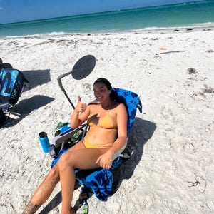 May include: A woman in a yellow bikini sits in a blue beach chair on a white sandy beach. She is giving a thumbs up. The chair is positioned near the ocean, with a blue water bottle and a black sun reflector nearby.