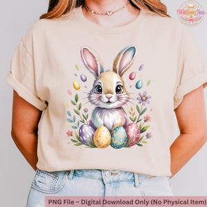 May include: Beige t-shirt with a watercolour illustration of a bunny surrounded by Easter eggs and flowers. The bunny has pink and blue accents. The text at the bottom reads "PNG File - Digital Download Only (No Physical Item)".