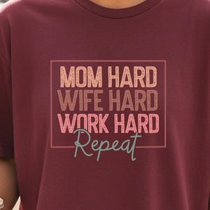 May include: Burgundy crew neck t-shirt with the text "MOM HARD WIFE HARD WORK HARD Repeat" in a rectangular box. The shirt is worn by a person holding a clear plastic cup with a brown beverage.