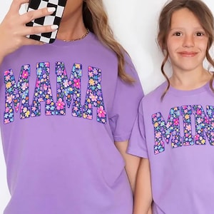 May include: Two matching lavender t-shirts, one adult and one child size, with the word "MAMA" and "MINI" in floral print. The adult is wearing a baseball cap and black shorts. The child is wearing black shorts. Both are holding hands.