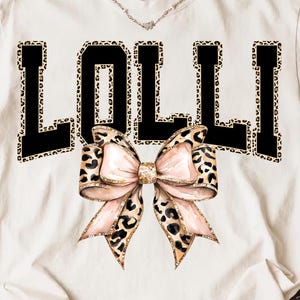 Könnte beinhalten: Weißes T-Shirt mit dem Wort "LOLLI" in schwarzen Blockbuchstaben mit Leopardenmuster. Eine große Schleife mit rosa und Leopardenmuster-Akzenten ist unter dem Text zentriert. Eine silberne Halskette ist sichtbar.