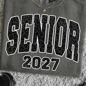 May include: A charcoal gray crewneck t-shirt featuring the word SENIOR in large, block-style letters with a black leopard print pattern and a white outline, followed by the number 2027 in the same leopard print style with a white outline.