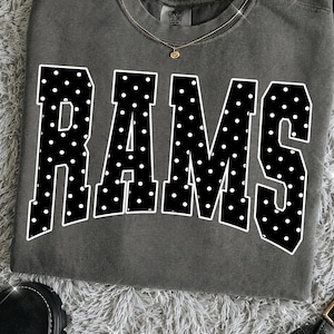 May include: A charcoal gray crewneck t-shirt featuring the word RAMS in large, bold, black block letters with a white polka dot pattern. The shirt has a visible Comfort Colors brand tag at the inner neckline and a thin gold chain necklace.