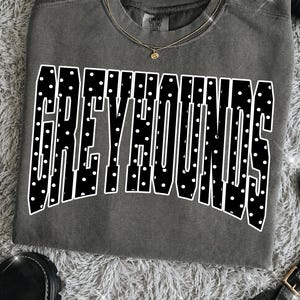 May include: A charcoal gray crewneck t-shirt featuring the word GREYHOUNDS in large, bold, black block letters with a white polka dot pattern. The shirt has a classic fit with a ribbed neckline and a visible brand tag inside the collar.