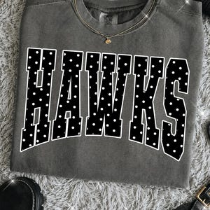 May include: A charcoal gray crewneck t-shirt featuring the word HAWKS in large, bold, black block letters with a white polka dot pattern. The shirt has a ribbed neckline and a Comfort Colors brand label visible at the inner collar area.