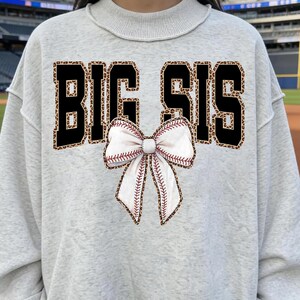 Puede incluir: Una sudadera gris claro con el texto "BIG SIS" en letras negras con un estampado de leopardo. Un lazo blanco con costuras rojas, que se asemeja a una pelota de béisbol, está centrado debajo del texto. La sudadera tiene cuello redondo y dobladillo con volantes.
