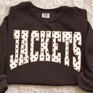 May include: A dark brown long-sleeve sweatshirt featuring the word JACKETS in large, cream-colored block letters with a black polka dot pattern. The text is arched across the chest, and the garment has a crew neckline with a Comfort Colors label.
