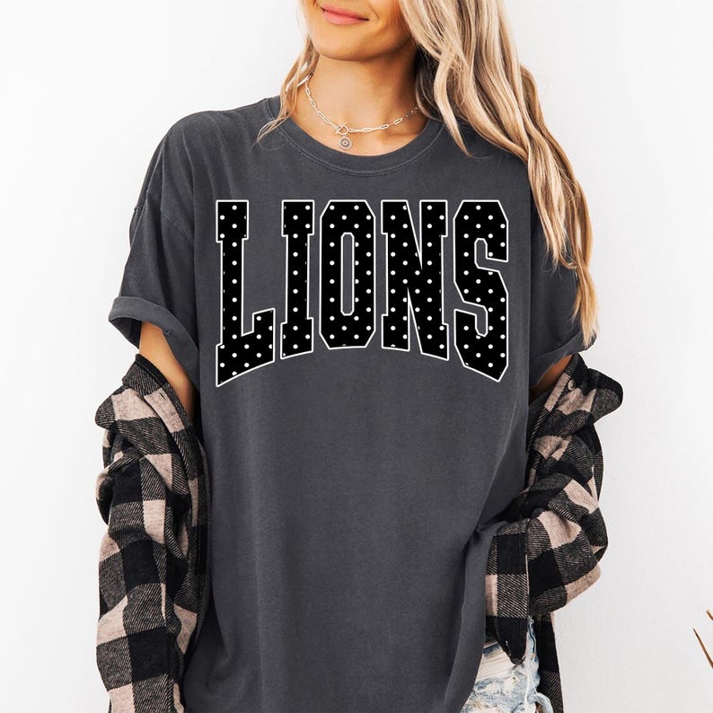 May include: A charcoal gray short-sleeved t-shirt featuring the word LIONS in large, block-style letters across the chest. The letters are black with a white polka dot pattern and a white outline, arranged in a slight arch across the front.