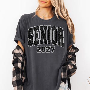 May include: A charcoal gray short-sleeve t-shirt featuring the word SENIOR in a black leopard print pattern with a white outline, followed by the year 2027 in white text. The shirt is worn with a black and beige buffalo plaid flannel shirt.