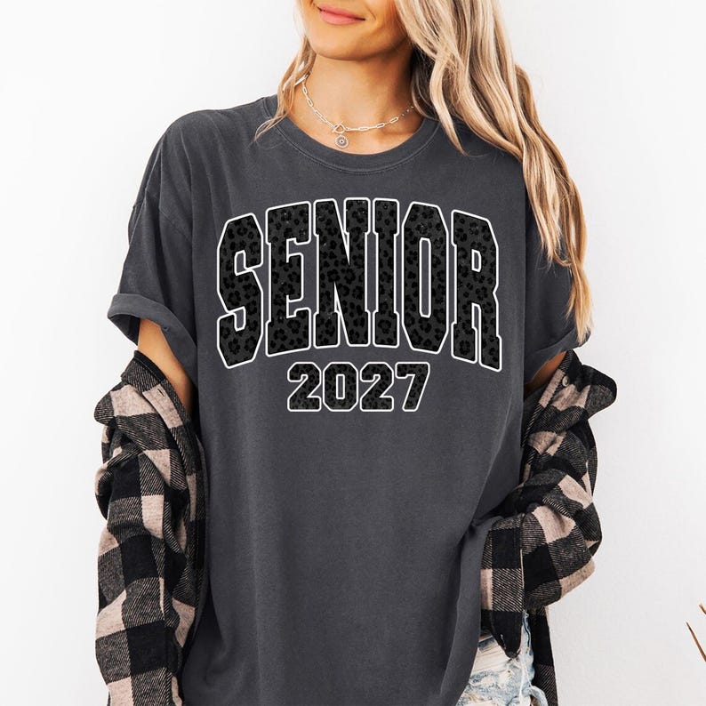 May include: A charcoal gray short-sleeve t-shirt featuring the word SENIOR in a black leopard print pattern with a white outline, followed by the year 2027 in white text. The shirt is worn with a black and beige buffalo plaid flannel shirt.