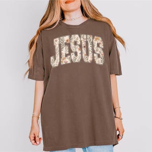 Peut inclure: T-shirt marron avec le mot "JESUS" en grandes lettres fleuries sur la poitrine. Les lettres sont de couleur cr&egrave;me et les fleurs sont dans les tons de rose, d'orange et de vert. Le t-shirt a des manches courtes et une coupe ample.