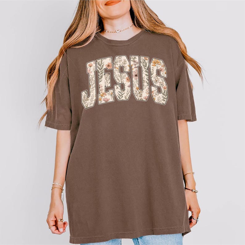 Peut inclure: T-shirt marron avec le mot "JESUS" en grandes lettres fleuries sur la poitrine. Les lettres sont de couleur cr&egrave;me et les fleurs sont dans les tons de rose, d'orange et de vert. Le t-shirt a des manches courtes et une coupe ample.