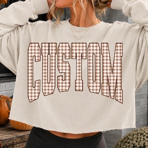May include: A beige cropped sweatshirt featuring the word CUSTOM printed in a brown and white gingham check pattern. The sweatshirt has a crew neckline, long sleeves with ribbed cuffs, and a raw, unfinished hemline at the waist.