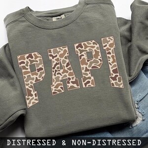 May include: An olive green sweatshirt featuring the word "PAPI" in large, block letters. The letters are filled with a camouflage pattern in shades of brown and tan. The text "DISTRESSED & NON-DISTRESSED" is at the bottom.