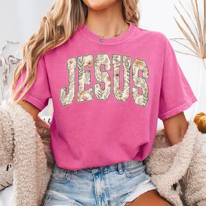 Peut inclure: T-shirt rose vif &agrave; manches courtes avec le mot &laquo; JESUS &raquo; imprim&eacute; sur le devant dans un motif floral. Les lettres sont remplies d'un fond cr&egrave;me et d'un motif de fleurs et de feuilles.