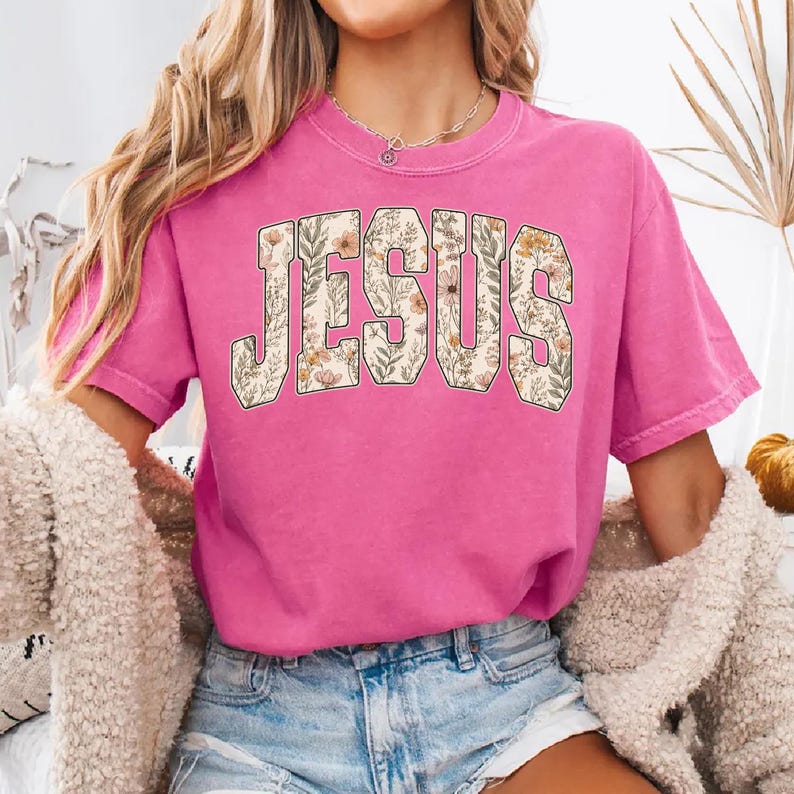 Peut inclure: T-shirt rose vif &agrave; manches courtes avec le mot &laquo; JESUS &raquo; imprim&eacute; sur le devant dans un motif floral. Les lettres sont remplies d'un fond cr&egrave;me et d'un motif de fleurs et de feuilles.