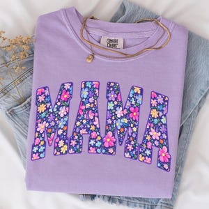 May include: A lilac Comfort Colors sweatshirt with the word "MAMA" in large, floral-filled letters. A gold necklace with a small pendant rests on the shirt, which is folded on top of a pair of blue jeans.