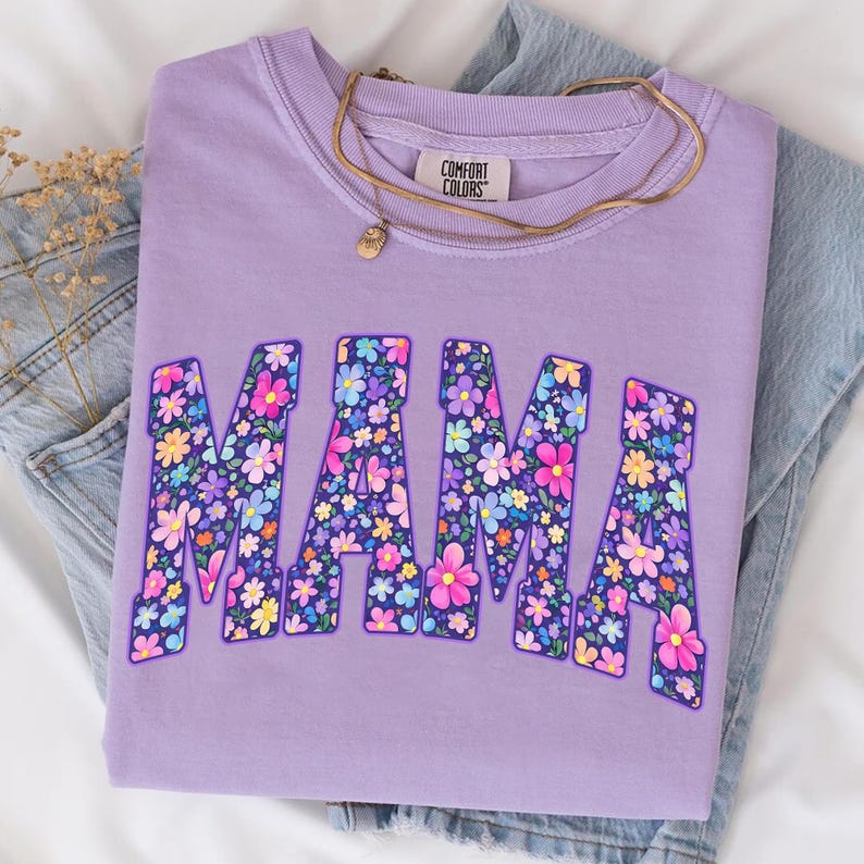 May include: A lilac Comfort Colors sweatshirt with the word "MAMA" in large, floral-filled letters. A gold necklace with a small pendant rests on the shirt, which is folded on top of a pair of blue jeans.