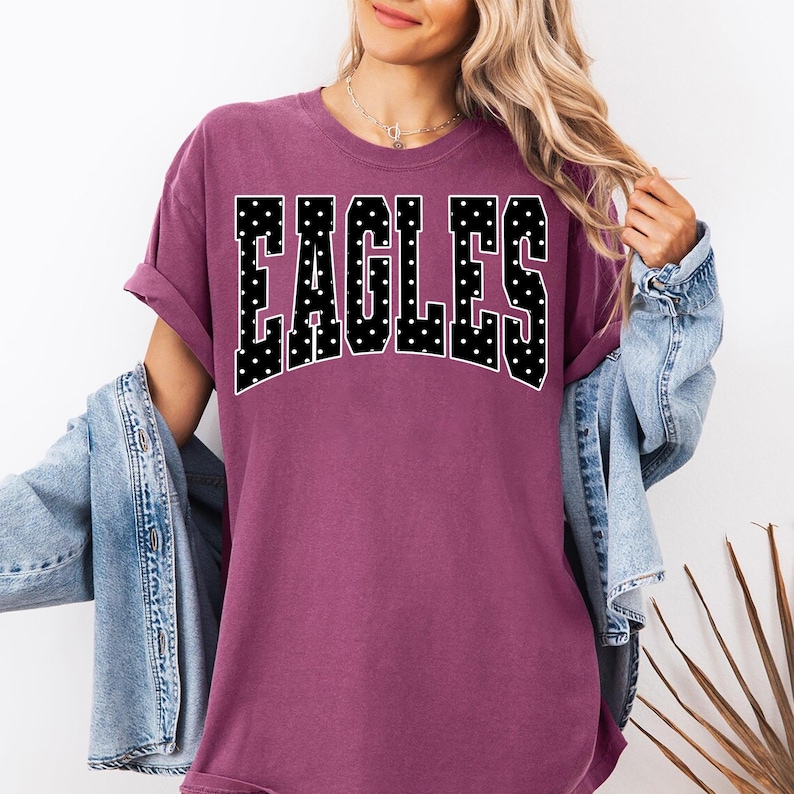 May include: A plum-colored short-sleeve t-shirt featuring the word EAGLES printed in a large, arched, black block font with white polka dots. The shirt has a crew neckline and a relaxed fit, paired with a light blue denim jacket worn over it.
