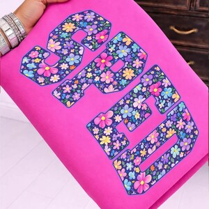 Pode incluir: Tecido rosa vibrante com a palavra "SPED" num padr&atilde;o floral. As letras s&atilde;o contornadas em azul escuro e preenchidas com flores coloridas em tons de rosa, amarelo, azul e roxo. O tecido &eacute; segurado por uma m&atilde;o.