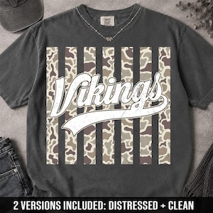 May include: A charcoal gray short-sleeve t-shirt featuring a central graphic of vertical stripes with a camouflage pattern. The word Vikings is printed in a large, white, distressed script font across the center. The shirt has a crew neckline.