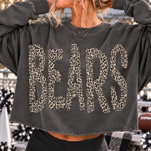 May include: A charcoal gray long-sleeve sweatshirt with a raw-edge hem. The front features the word BEARS in large, bold capital letters filled with a tan and black leopard print pattern. The fabric has a faded, vintage-style wash throughout.