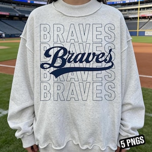 May include: A light gray sweatshirt with the word "BRAVES" in navy blue, stacked vertically. The word "Braves" is also written in a cursive font in the center. The sweatshirt has a crew neck and a wavy hemline. The background shows a baseball field.