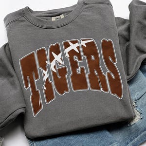 May include: A charcoal gray crewneck sweatshirt featuring the word TIGERS in large, brown, block-style letters. The letters have a white outline and a white graphic pattern resembling the laces of an American football across the center.