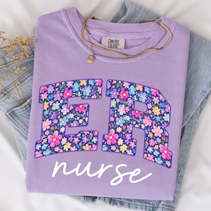May include: A lilac Comfort Colors sweatshirt with the text "ER nurse" in white script. The letters "ER" are filled with a colorful floral pattern. A gold necklace with a pendant rests on the shirt, which is folded on top of blue denim.