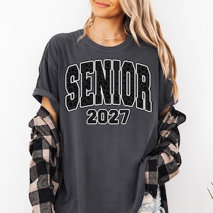 May include: A charcoal gray short-sleeve t-shirt featuring the word SENIOR in a black leopard print font with a white outline, followed by the number 2027 in a matching black leopard print font with a white outline, layered under a plaid shirt.