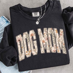Op de afbeelding: Zwart sweatshirt met de tekst "DOG MOM" in een bloemenpatroon. De crew neck sweatshirt heeft lange mouwen. Een zilveren ketting met een ronde hanger ligt op het kledingstuk.