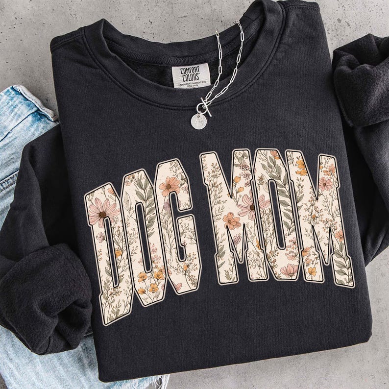 Op de afbeelding: Zwart sweatshirt met de tekst "DOG MOM" in een bloemenpatroon. De crew neck sweatshirt heeft lange mouwen. Een zilveren ketting met een ronde hanger ligt op het kledingstuk.