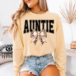 May include: A light yellow long-sleeve sweatshirt with the word "AUNTIE" in black leopard print letters. A pink and leopard print bow is centered below the word. The sweatshirt is paired with denim shorts.