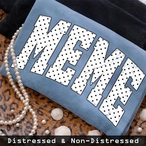 May include: A light blue t-shirt with the word "MEME" in large, distressed white letters with black polka dots. The shirt is folded and resting on a leopard print surface, with a pearl necklace and seashells.
