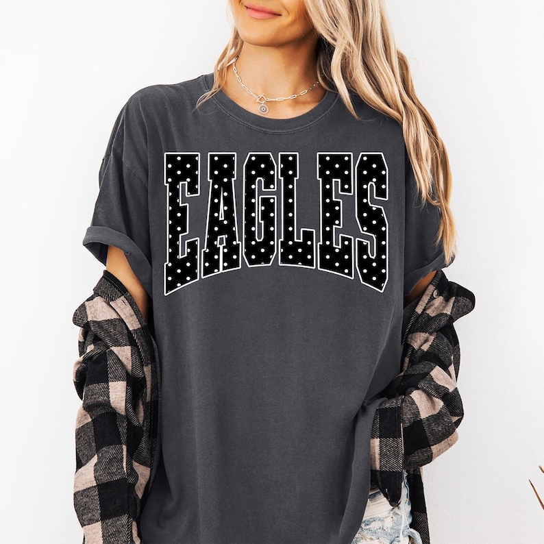 May include: A charcoal gray short-sleeve t-shirt featuring the word EAGLES in a large, arched, collegiate-style font. The letters are black with a white polka dot pattern and a thin white outline. A black and tan plaid flannel shirt is layered.