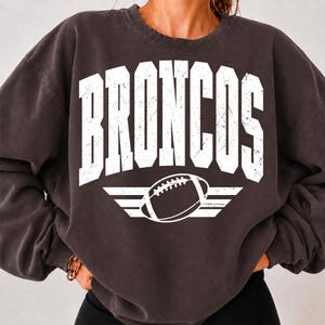 Puede incluir: Una sudadera de cuello redondo en color gris carbón que presenta la palabra BRONCOS en letras mayúsculas blancas grandes y desgastadas arqueadas sobre el pecho, sobre un gráfico de un balón de fútbol americano con alas horizontales en blanco.