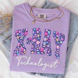 May include: A lilac Comfort Colors sweatshirt with the words "X-RAY Technologist" in a floral design. The letters are filled with colorful flowers. A gold necklace rests on the shirt, which is folded on top of a pair of blue jeans.