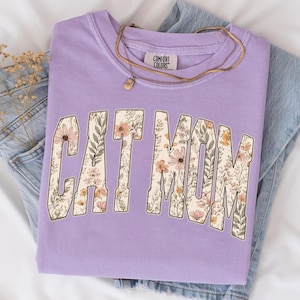 May include: A lilac sweatshirt featuring the text "CAT MOM" filled with a floral pattern. The crew neck sweatshirt has long sleeves. A gold necklace with a pendant is placed on the shirt. The tag reads "COMFORT COLORS".