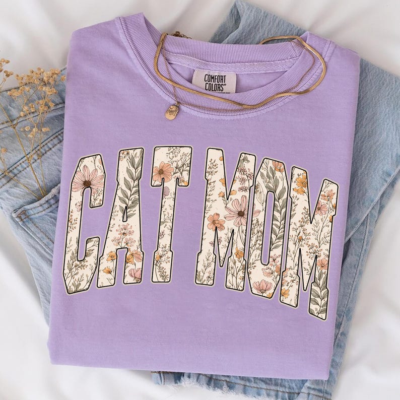 May include: A lilac sweatshirt featuring the text "CAT MOM" filled with a floral pattern. The crew neck sweatshirt has long sleeves. A gold necklace with a pendant is placed on the shirt. The tag reads "COMFORT COLORS".