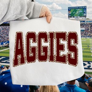 May include: White rectangular textile with the word "AGGIES" in maroon letters with a leopard print border. The textile is held up in front of a football stadium.